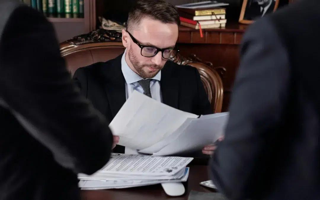 A man in glasses and a suit examines documents at a desk cluttered with papers while two others stand nearby, perhaps discussing the nuances of apostille vs notary services. Books and office items fill the background, reinforcing the professional setting.