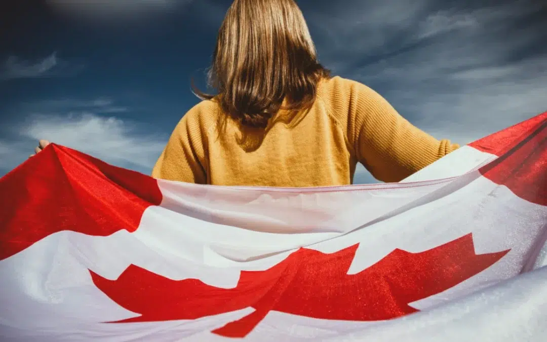 A person with long hair stands facing a blue sky, wrapped in a large Canadian flag. They wear a mustard-colored sweater, and the flag's red maple leaf is prominently visible—a serene moment reminiscent of the global unity found in agreements like the Hague Apostille Convention.