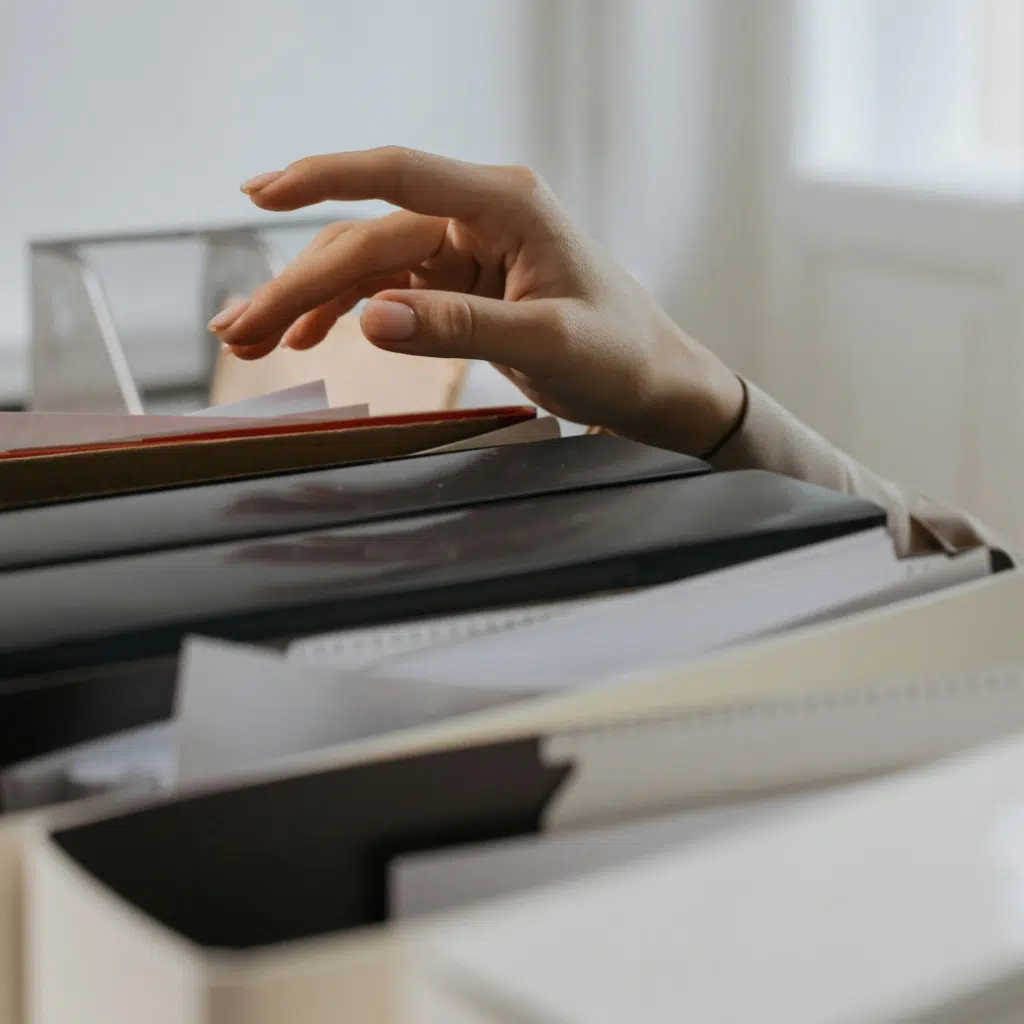 A hand reaching into a stack of folders and papers, with fingers gently pulling out a birth certificate for apostille. The background is softly blurred, suggesting an office or workspace setting.