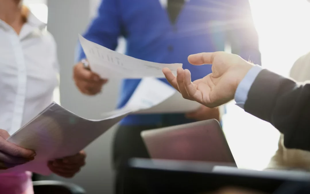 In an office bathed in sunlight, a group of professionals exchange documents and discuss topics like how to get a Canadian birth certificate. Some hold papers while others focus on their laptops, fully engaged in the collaborative atmosphere.