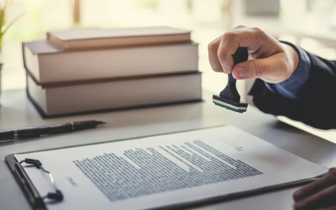 A person stamps a document on a clipboard, perhaps processing a Canadian death certificate. The scene features a stack of books in the background, with the tabletop brightly lit by natural light. The focused hand holding the stamp suggests an official or administrative setting.