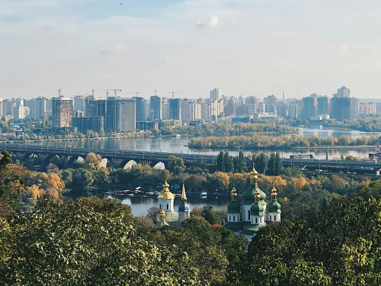 A scenic view of a city with a river flowing through it. A large bridge spans the river, and tall buildings line the skyline. In the foreground, a cluster of green-domed churches peeks through lush trees, surrounded by autumn foliage.