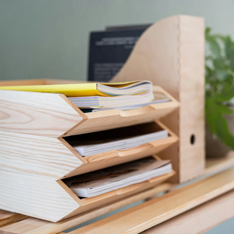 A wooden desk organizer holds several magazines and papers in its three-tier slots. A green plant is partially visible in the background, adding a touch of nature to the tidy workspace.