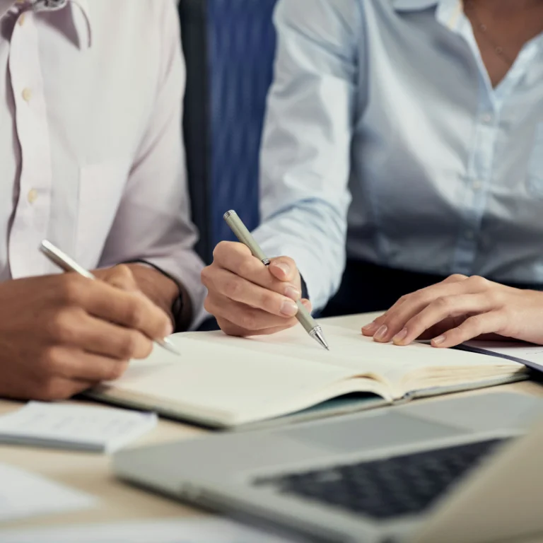 Two people seated at a table, taking notes in notebooks. They are using pens and wearing professional attire. A laptop and several papers are scattered on the table, indicating a work or study setting.