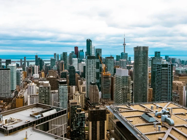 Aerial view of an Ontario cityscape featuring numerous skyscrapers under a cloudy sky. A prominent tower with a slender antenna stands amid the buildings. Rooftops and urban structures fill the foreground, creating a dense architectural scene that captures the essence of urban authenticity.