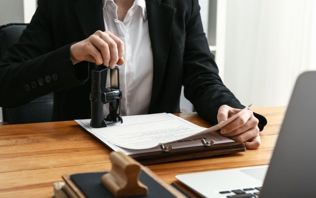 A person in a black suit uses a stamp on a document placed on a clipboard. The scene is set at a wooden desk with a laptop, additional stamp, and office supplies visible.