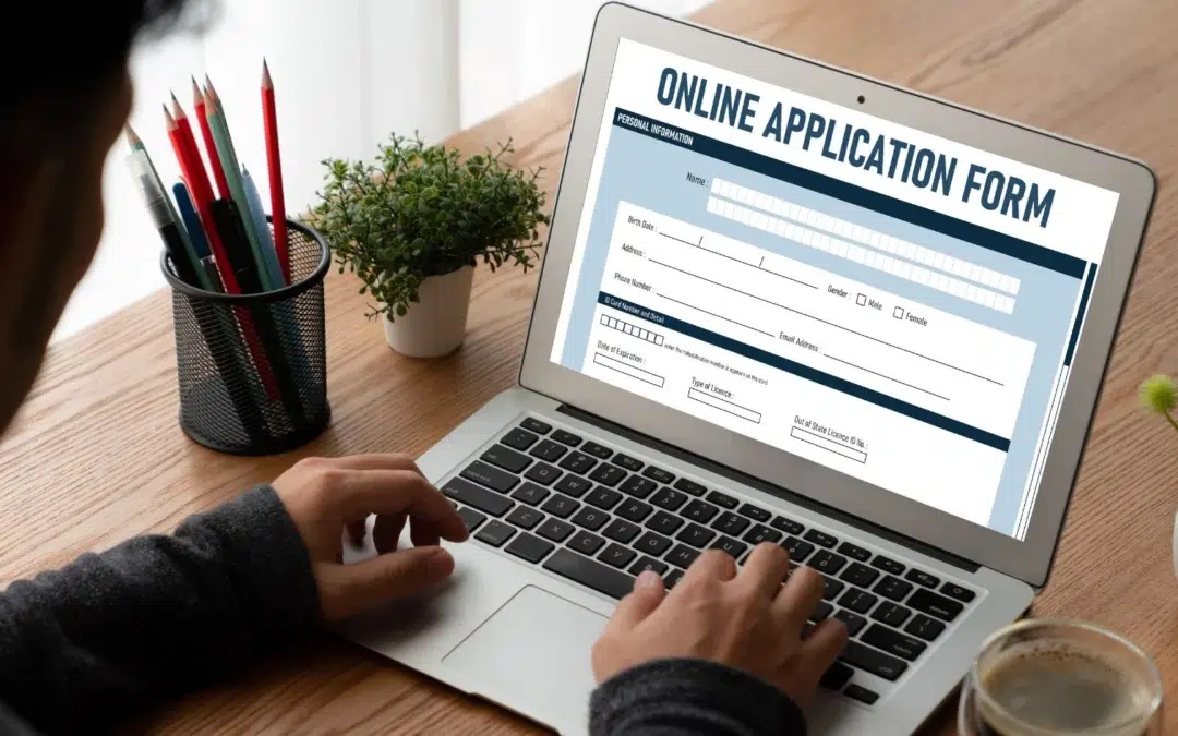 A person is sitting at a wooden desk, typing on a laptop displaying an online application form. Nearby are a small plant and a cup holding colored pencils. The form appears mostly blank, with fields for Name, Date of Birth, and Contact Information.