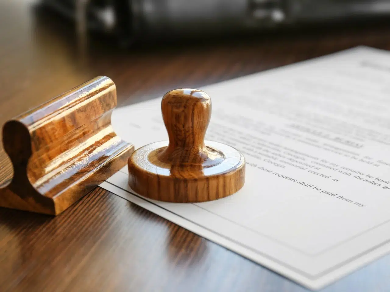 A wooden stamp and its holder rest on a document placed on a wooden table. The document appears to be a formal letter or contract, partially visible and printed in black text. Natural light illuminates the scene.