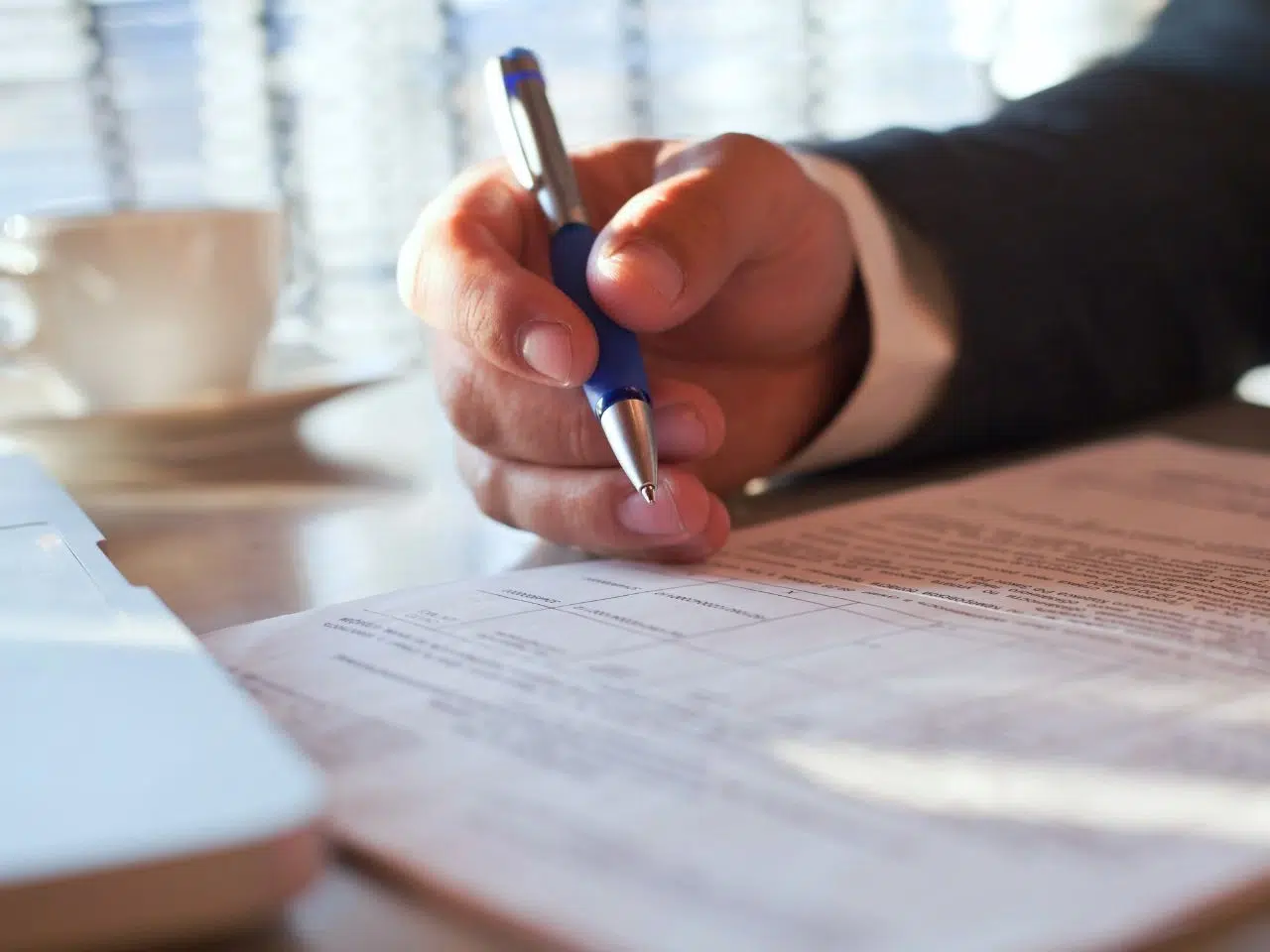 A person in a suit is holding a blue pen, pointing at a document on a desk. There is a laptop partially visible on the left and a blurred coffee cup in the background. The scene suggests a professional or business setting.