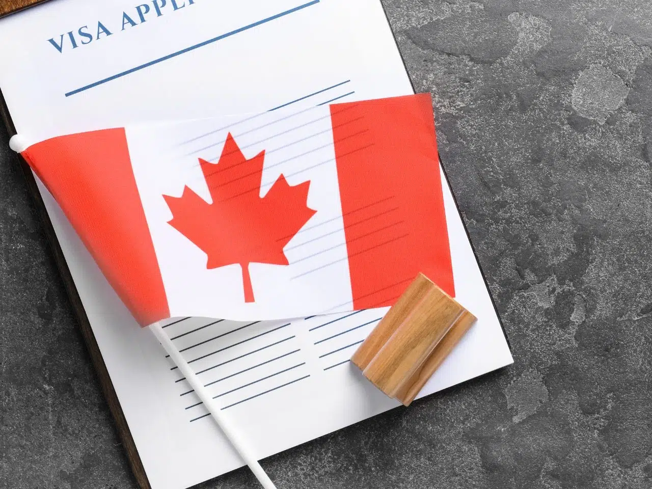 A Canadian flag lies on top of a visa application form on a textured dark surface. A wooden stamp is positioned nearby.
