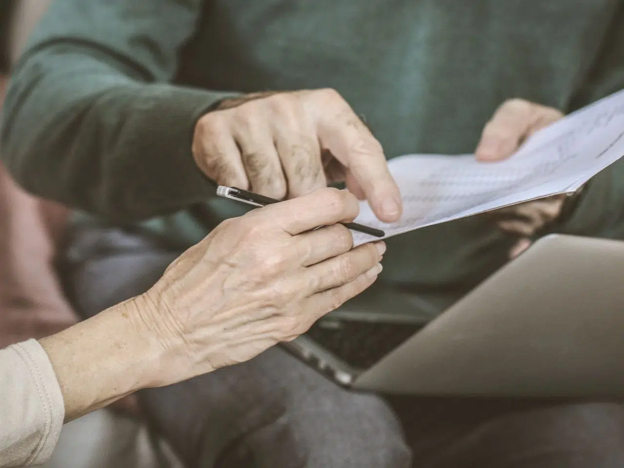 Two people are sitting with a laptop. One person is holding and pointing at a document, while the other holds a pen. They appear to be discussing or reviewing the contents of the paper.