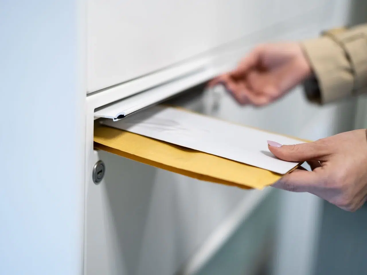 A person placing an envelope and a brown padded envelope into a mailbox slot. The image focuses on the hands and the items being mailed, with a neutral background.