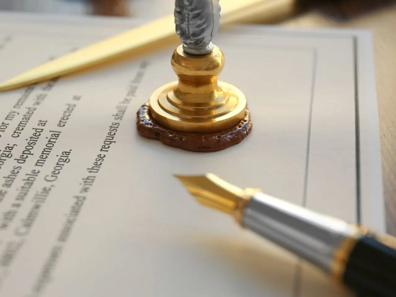 A fountain pen rests on a scroll of paper next to a golden sealing stamp on a wax seal. The focus is on the pen nib and the stamp, with the papers text blurred in the background.