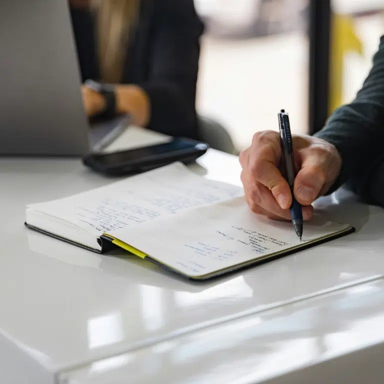 A person writes in a notebook on a white table with a pen in their right hand. A smartphone and laptop are nearby. Another person works on the laptop in the background. The scene suggests a work or study environment.