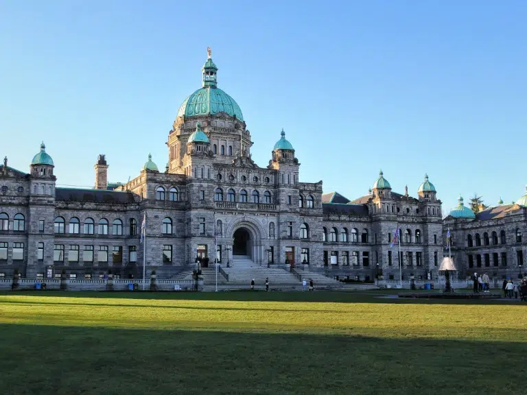 The image shows a large historic building with a central dome and multiple arches. The architecture features a blend of stone and ornate details, with a well-maintained lawn and a fountain in front. The sky is clear and blue.