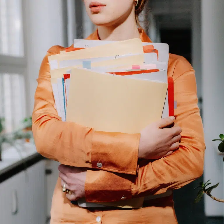 Two people are leaning over a table, signing documents. One person is wearing a watch and a red jacket, holding a pen, while the other persons hand rests on the papers. A bouquet of white flowers is visible in the foreground.