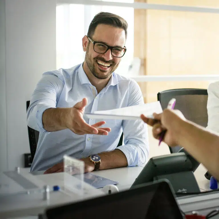 A smiling man in glasses reaches for a document handed to him by another person. He sits at a desk with digital tablets and paperwork, wearing a light blue shirt. The background shows a bright office space.