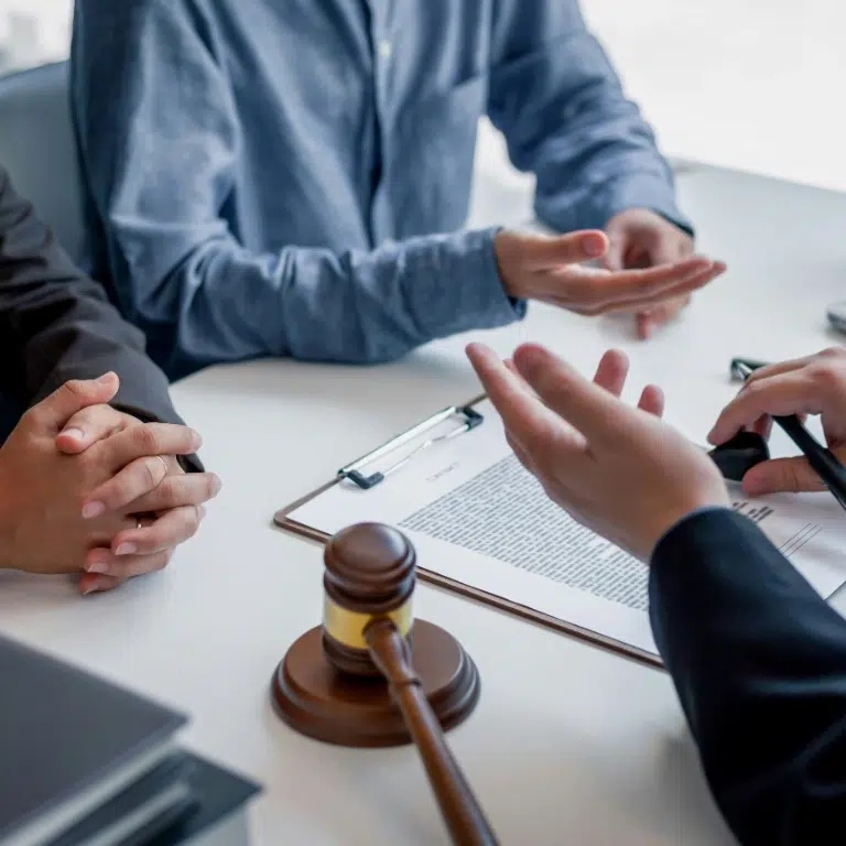 Three people are seated around a table. One person is holding a pen near a clipboard with documents. A gavel rests on the table, suggesting a legal or professional discussion. Hands are gesturing as if explaining or negotiating.