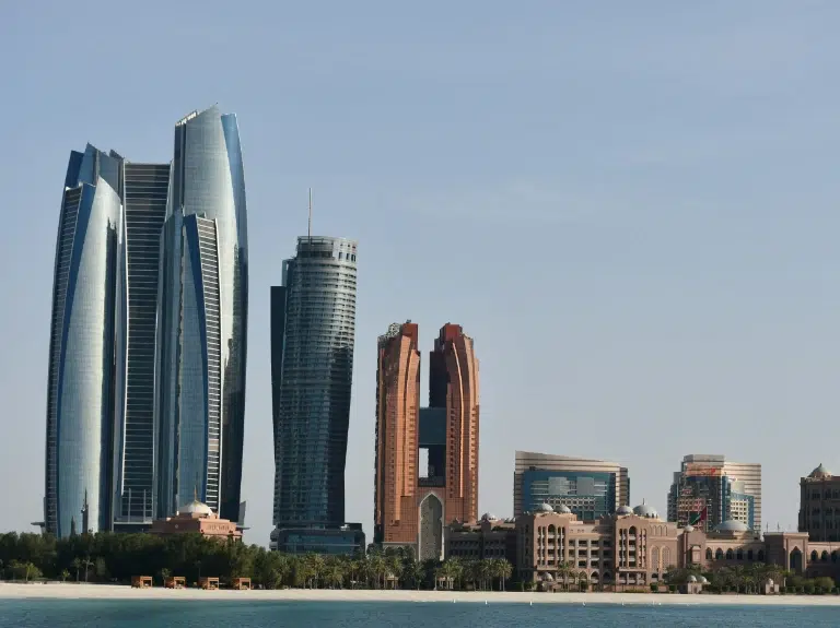 Skyline of Abu Dhabi featuring modern skyscrapers, including the distinctive Etihad Towers and other high-rise buildings, set against a clear blue sky. The buildings are situated by the waterfront with greenery in the foreground.
