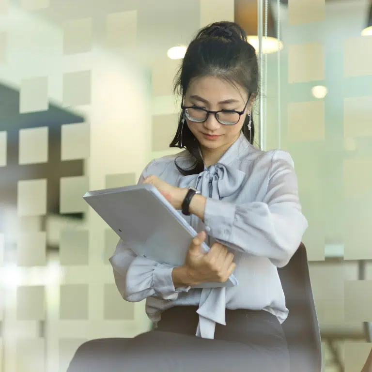 A woman in glasses sits on a chair, looking at her watch while holding a document. She wears a light blouse with a bow and has her hair tied back. The glass wall behind her has square patterns. The setting appears to be an office or meeting room.