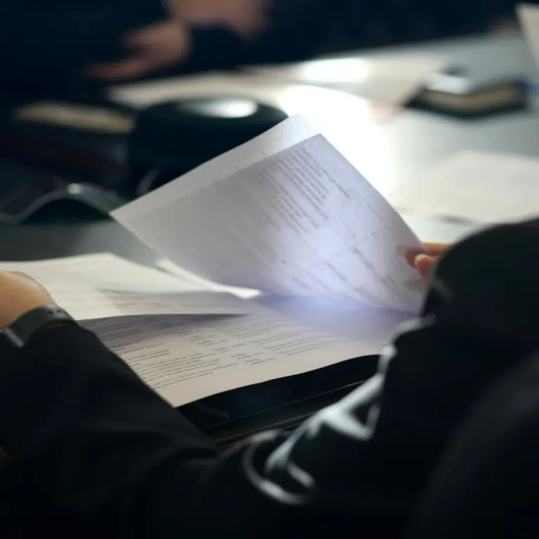 A person holding and reviewing several sheets of paper in a dimly lit office setting. The focus is on the documents, which are slightly blurred, while a blurry background suggests a meeting or work environment.