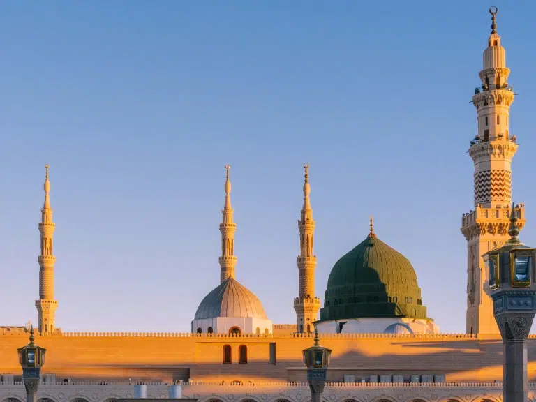 The image shows the Mosque of the Prophet in Medina, featuring its elegant architecture. Several minarets and a green dome are visible under a clear blue sky, illuminated by warm sunlight.
