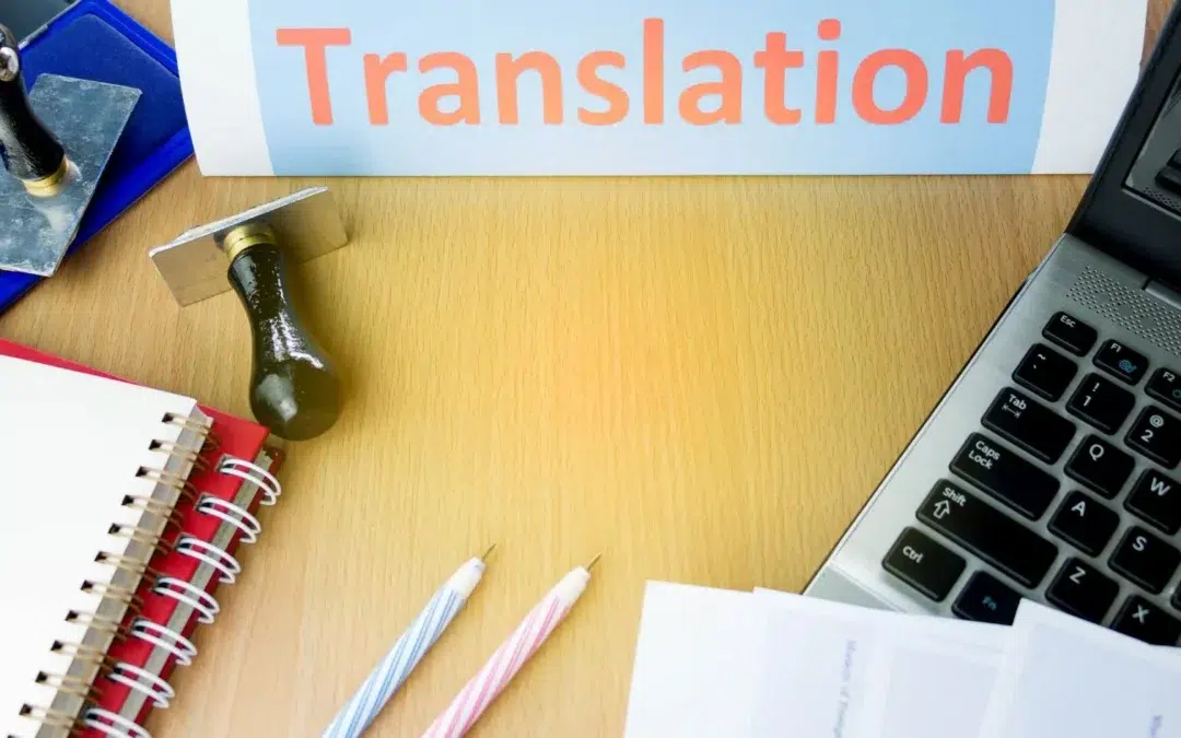 A desk with a laptop, a notebook, two pens, and ink stamps. A paper with the word Translation is seen on top. The scene is brightly lit, suggesting a work environment focused on translation tasks.