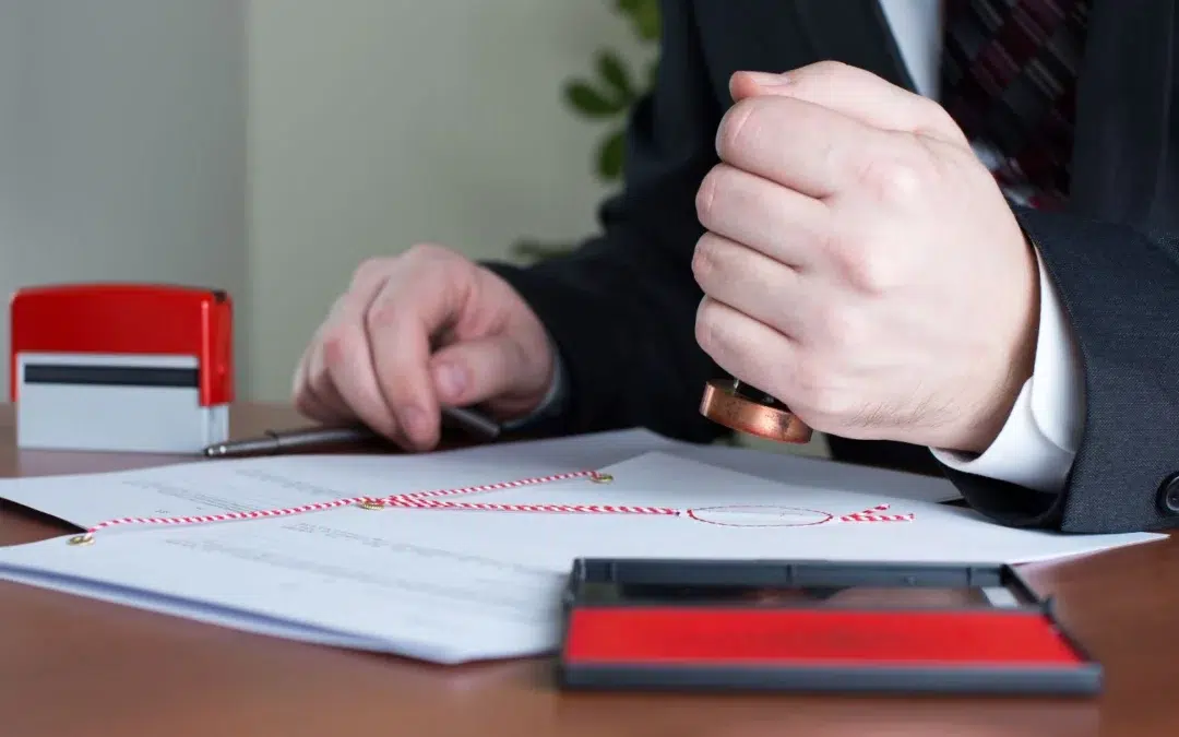 A person in a suit stamping a document on a desk. A red stamp pad is visible, and the paper features red and white twine. Another red stamp tool is on the table.