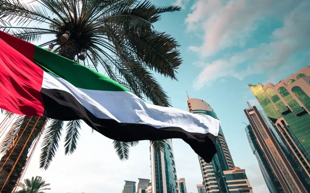 United Arab Emirates flag waving in front of modern skyscrapers and palm trees, under a partly cloudy sky.