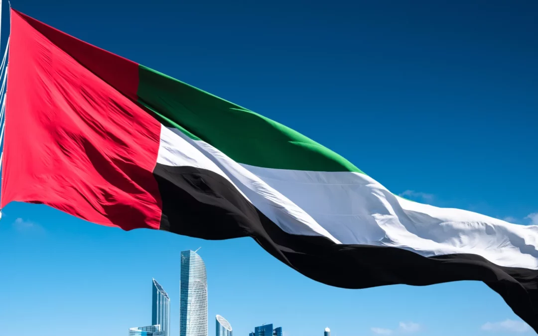 The flag of the United Arab Emirates waves in the wind against a clear blue sky, with modern skyscrapers visible in the background.