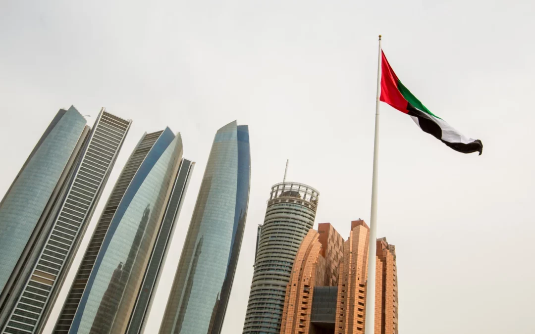 Several modern skyscrapers stand against a pale sky in Abu Dhabi, with the flag of the United Arab Emirates prominently flying on a tall flagpole in the foreground.