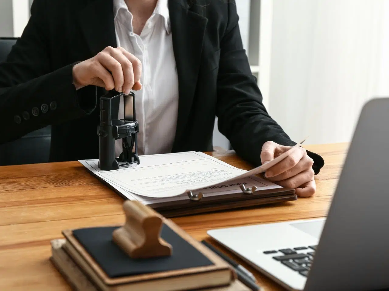 A person in a black suit is pressing a stamp onto a document on a wooden desk. Nearby are a clipboard, a rubber stamp, and a laptop. The scene suggests an office or formal setting, possibly involving document approval or verification.