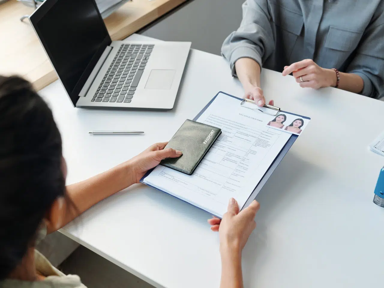 Two people sit at a desk with a laptop. One person hands a resume with photos and a passport to the other, suggesting a job interview or document verification process.