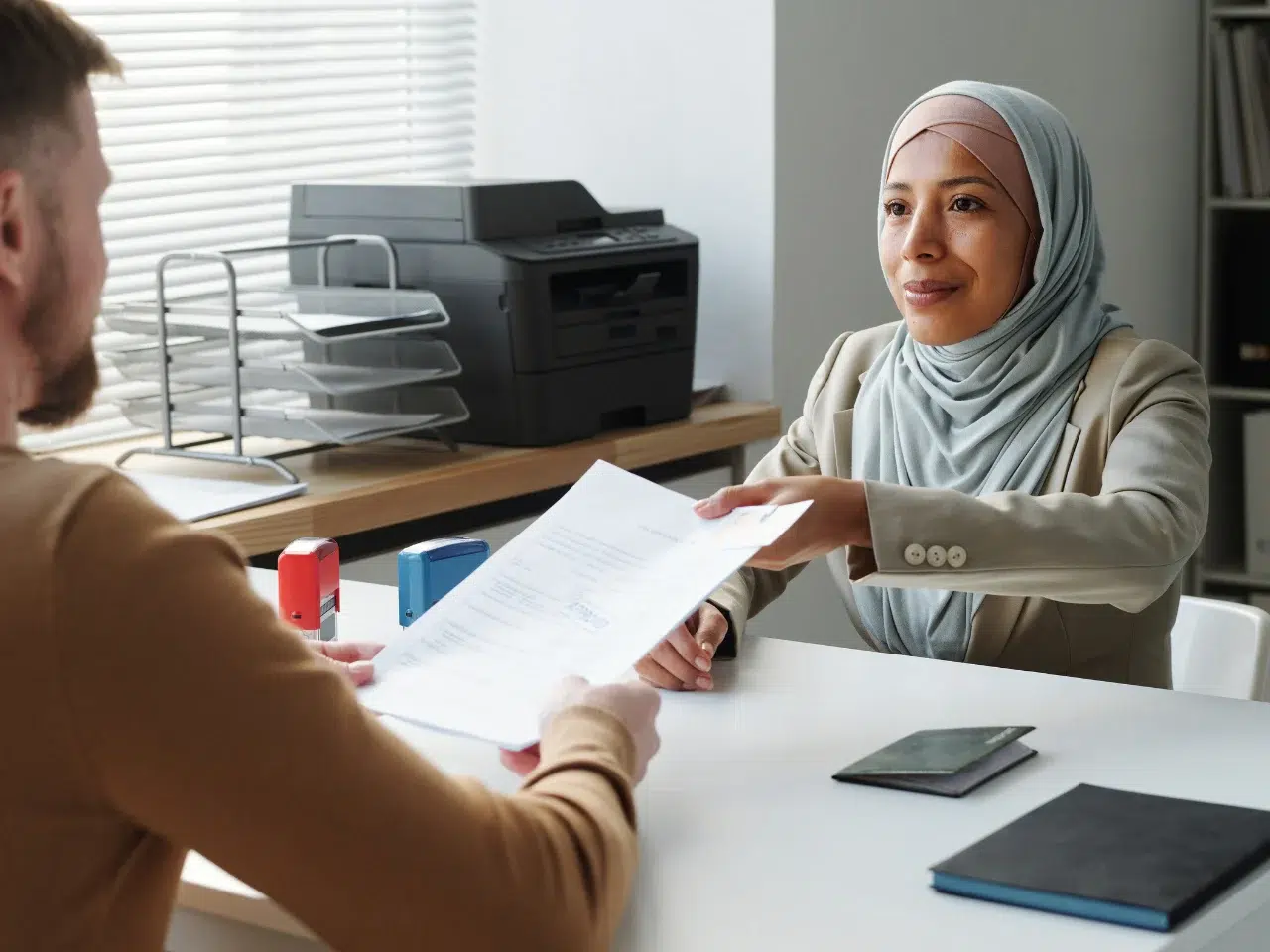 A woman in a hijab hands a document to a man across a desk in an office setting, with paperwork, notebooks, and a phone on the desk, and office equipment in the background.