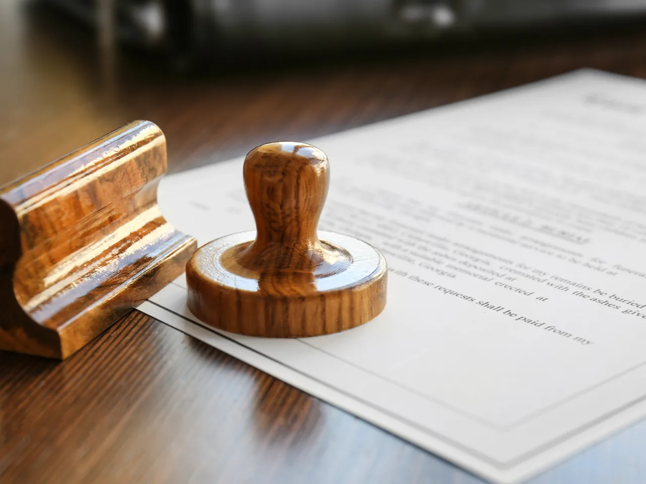 A wooden stamp and its holder rest on top of a document with printed text, placed on a wooden surface. The stamp appears ready to be used for certifying or approving the paper.
