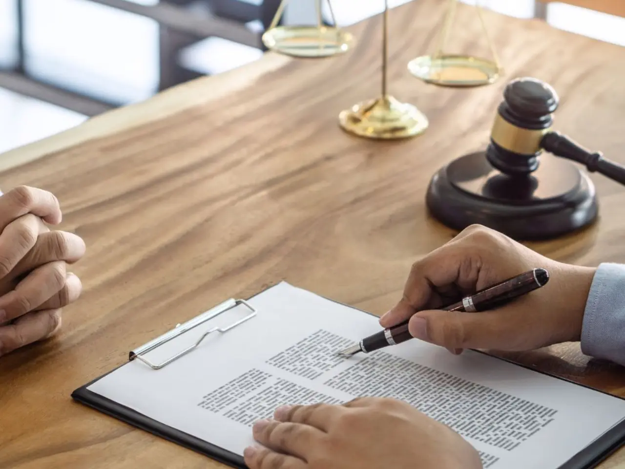 Two people sit at a wooden desk with legal documents on a clipboard, a pen in hand, and a judge’s gavel and scales of justice in the background, suggesting a legal or consultation setting.
