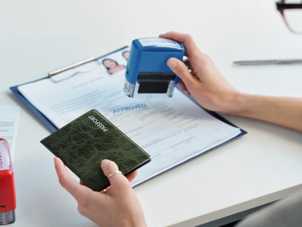 A person holds a green passport and uses a blue Approved stamp on a document clipped to a folder, with a pen and another red stamp visible on a white desk.