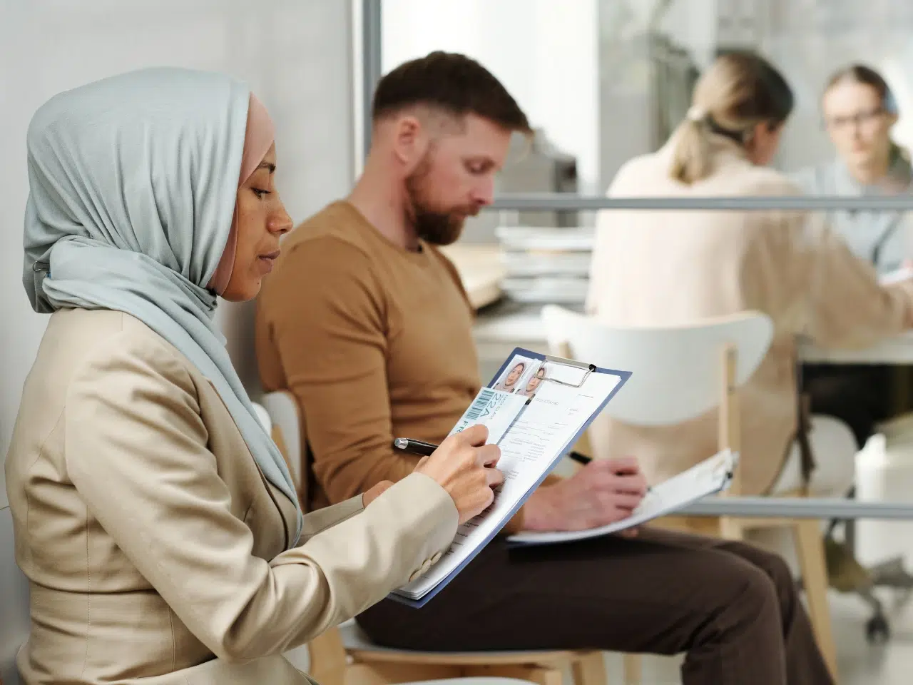 A woman in a light blue hijab and beige blazer fills out a form on a clipboard, while a man beside her writes on paper. Two blurred people are visible in the background through a glass divider.