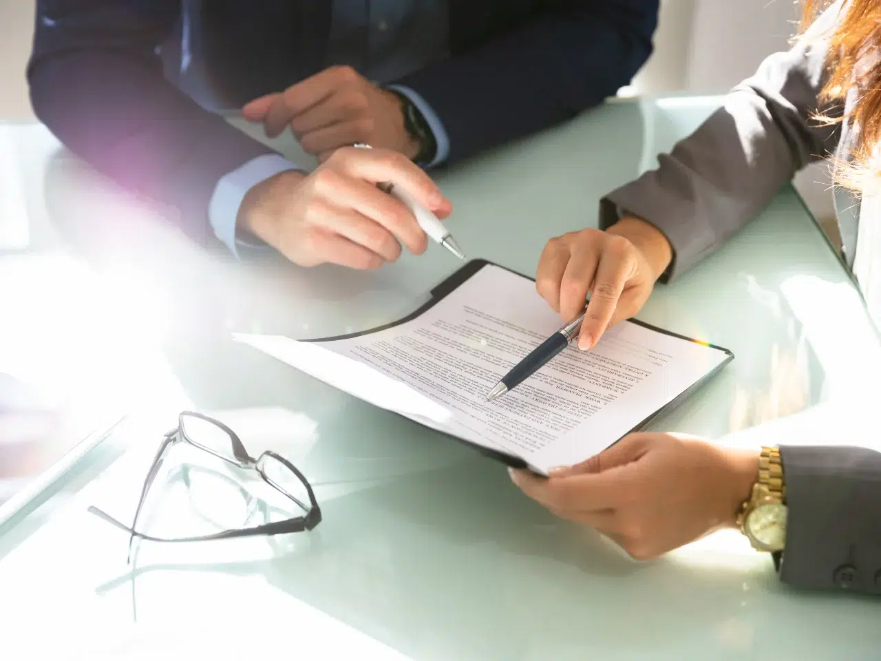 Two people in business attire review a document on a clipboard at a glass table, one pointing with a pen. A pair of eyeglasses rests nearby. Bright light shines from the left side of the image.