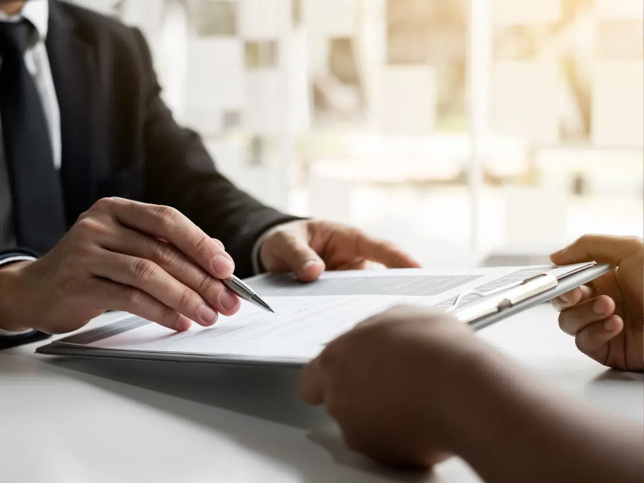 Two people in business attire sitting at a table, one holding a clipboard with documents while the other points at the paperwork with a pen, suggesting a discussion or signature process.