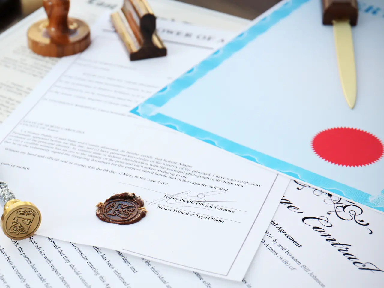 A collection of official documents, including a certificate with a blue border, paperwork, and a letter sealed with a wax stamp, are arranged on a desk with stamps and a gold letter opener.