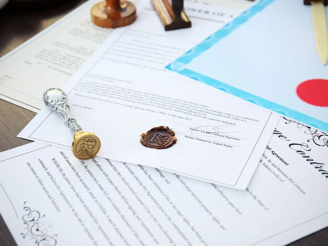 A collection of official documents and certificates on a desk, with embossed wax seals in gold and red. A shiny silver seal stamp is placed on top of the papers. The documents feature text and decorative borders.