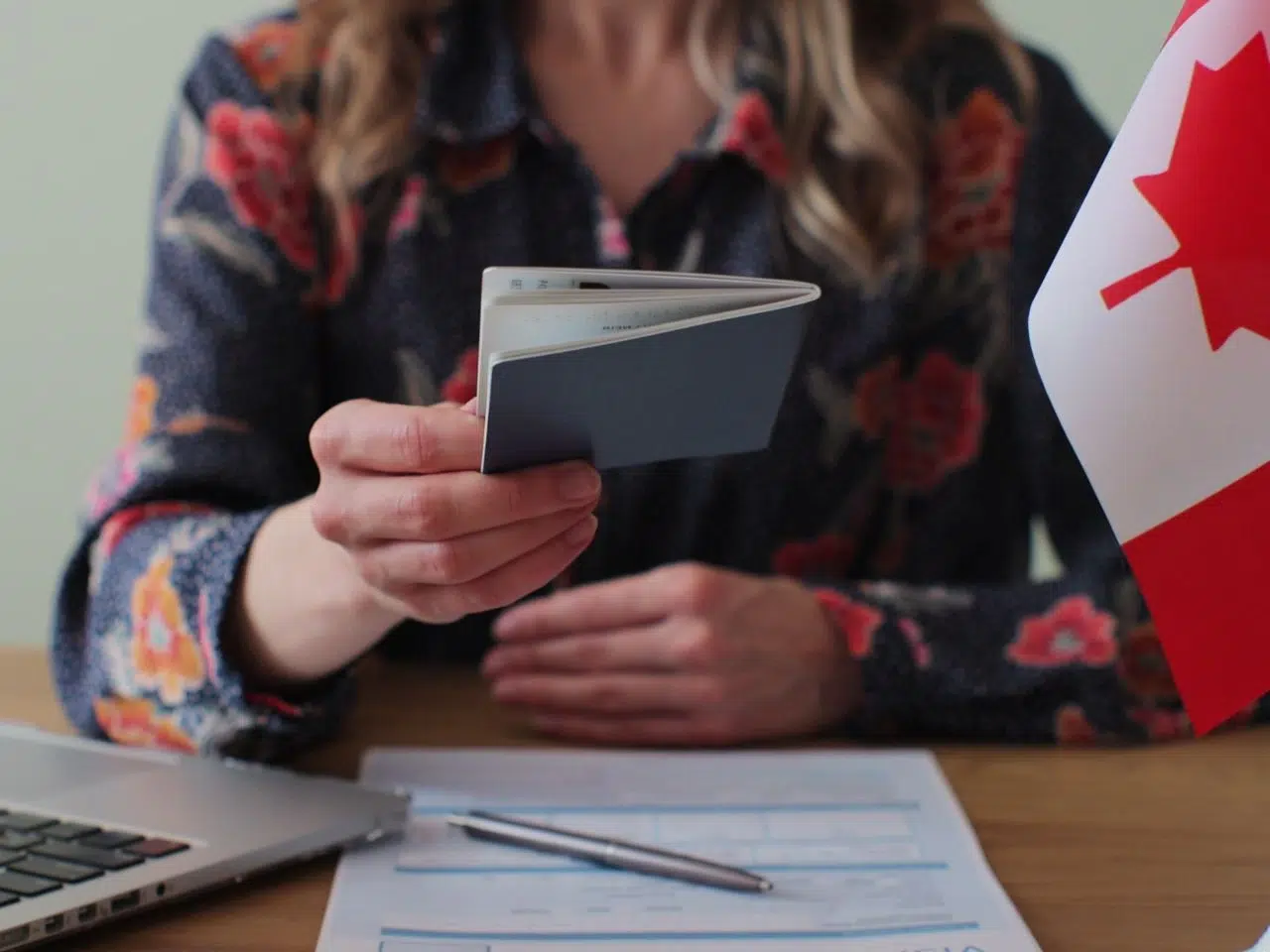 A person holding a passport sitting at a desk with a laptop and a form. A Canadian flag stands to the side. They are wearing a floral-patterned shirt. The scene suggests a travel or immigration context.