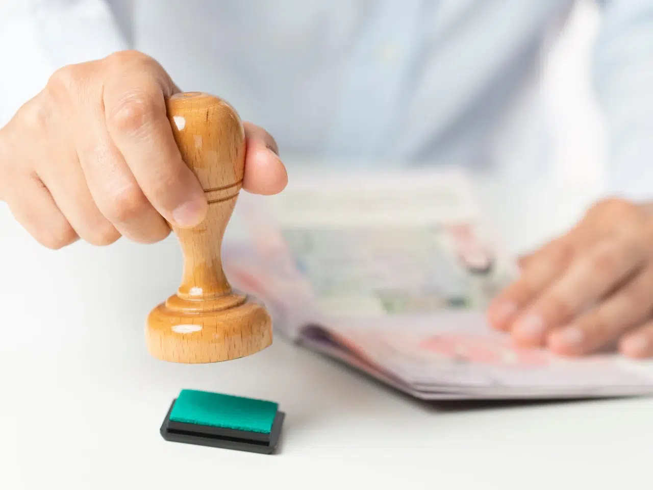 A person presses a wooden stamp onto a green ink pad, preparing to stamp a document or passport that is open on a white table.