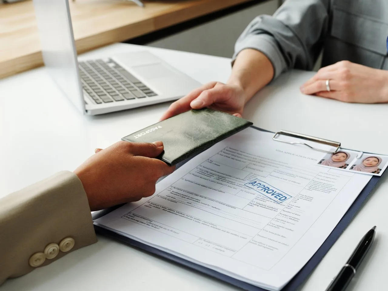 Two people exchange a passport over a desk with a laptop and an approved visa application form on a clipboard, along with two passport-sized photos.