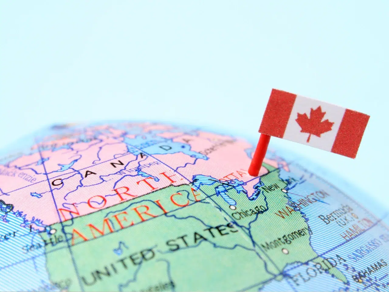A small Canadian flag is pinned on a globe, marking the country of Canada in North America. The map highlights Canada and parts of the United States with light blue and pink colors.