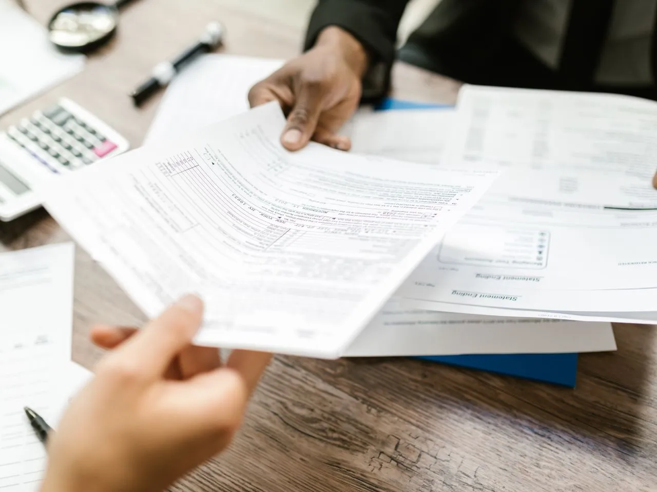 Two people are seated at a wooden table handling financial documents. One person is passing papers to the other. A calculator and a pen are visible on the table. The scene suggests a business meeting or financial discussion.