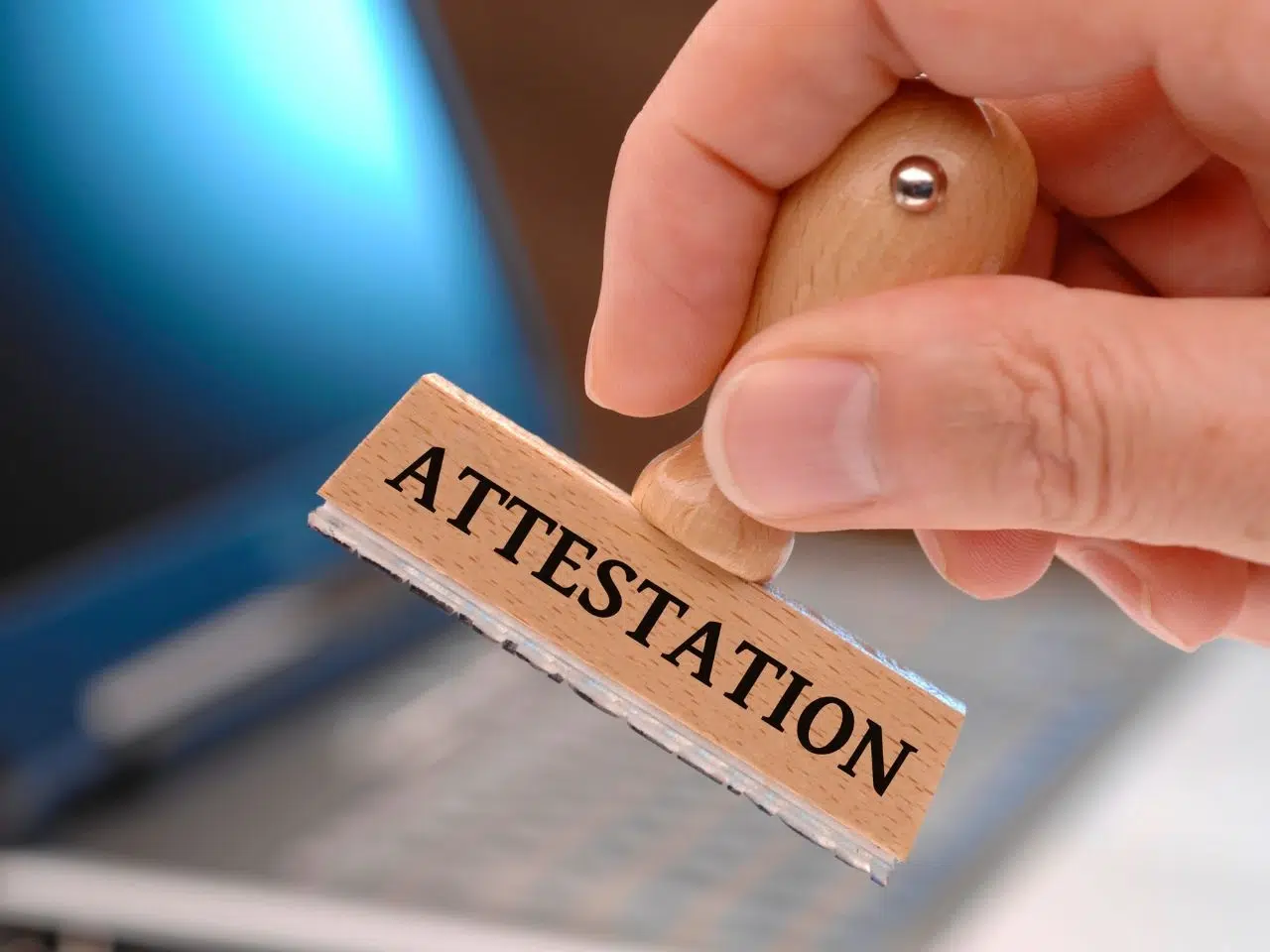 A hand holds a wooden stamp labeled ATTESTATION above a blurred laptop keyboard, suggesting the act of certifying or verifying documents in a digital context.