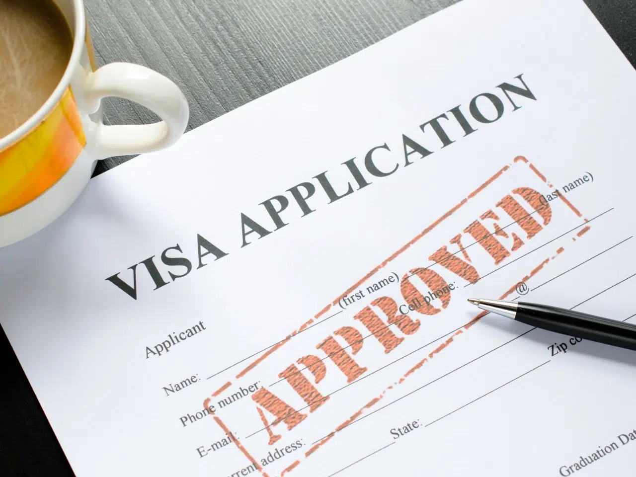 A visa application form with a large red APPROVED stamp across it, next to a pen and a cup of coffee on a dark table.