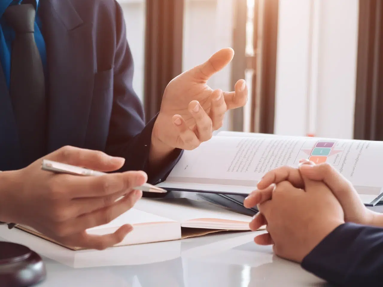 Two people in business attire sit at a desk, having a discussion. One person gestures with their hand while the other listens, hands clasped. An open book and documents are visible on the table.